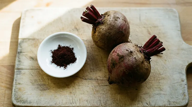 Two whole raw beets with soil on them beside a shallow dish of dark-red powder on a wooden board