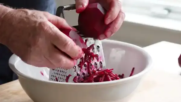 Hands grating a raw beet on a steel box grater over a white bowl, bright red shreds falling in