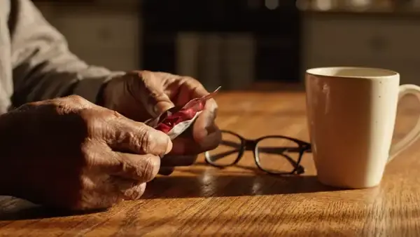 A man's hands turning a small dark-red pouch over on a kitchen table beside reading glasses and a mug